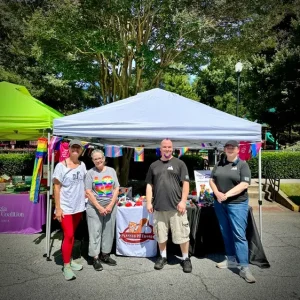 Four Planned PEThood of Georgia volunteers stand together in front of their white canopy tent decorated with pride flag bunting at their Gwinnett Pride 2026 booth in Norcross.